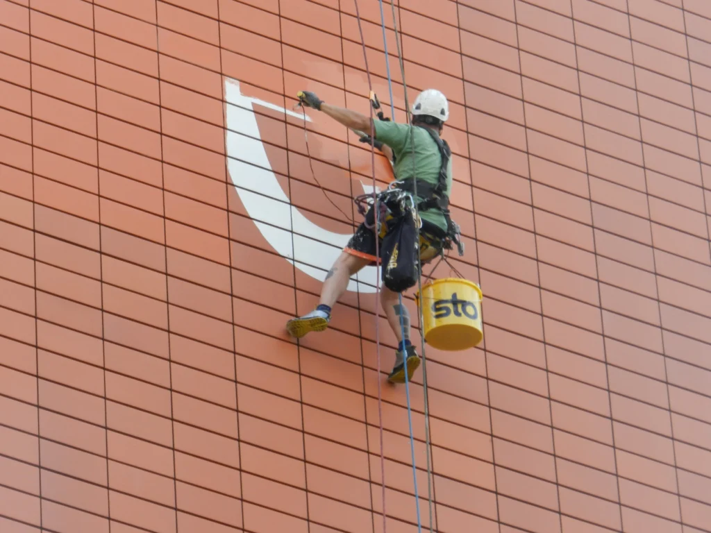 Person mit Helm und Sicherungsgurt arbeitet an einer roten Fassade mit Kletterausrüstung und Malerwerkzeug