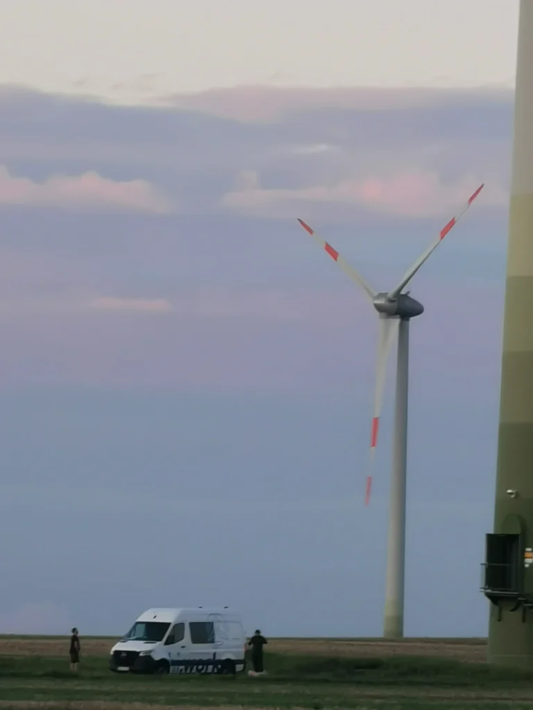 Windkraftanlage mit rot-weißen Rotorblättern auf offenem Feld, davor ein weißer Transporter und zwei Industriekletterern auf dem Boden
