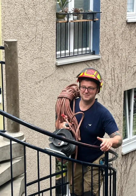 Ein Arbeiter mit Schutzhelm trägt eine große Rolle Seil und steht an einem Treppengeländer vor einem Haus mit Fenstern.